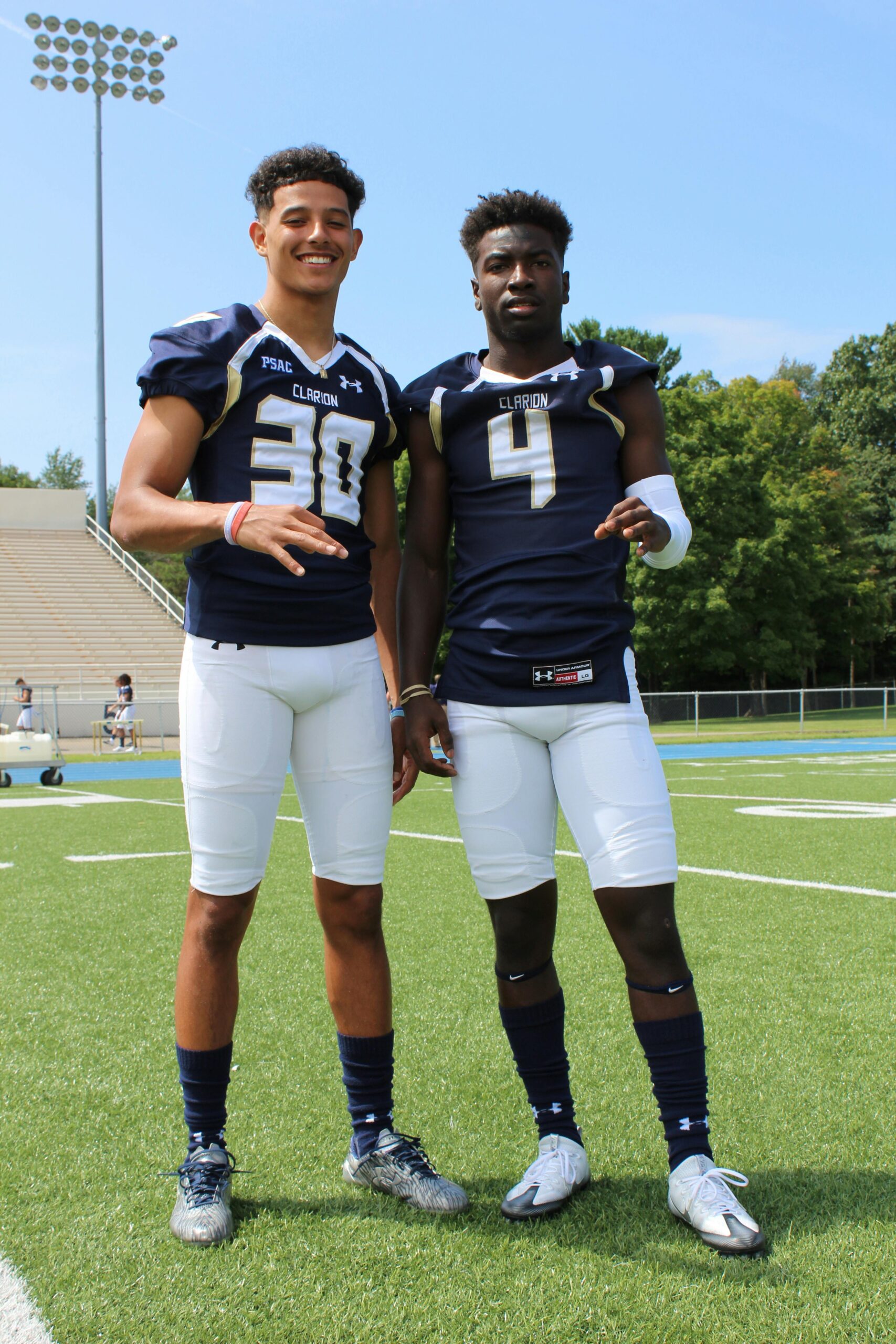 Two athletes in football uniforms pose on a sunny outdoor field.