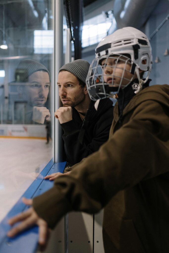 Father and son, in winter gear and hockey helmet, observing an ice hockey game intently.